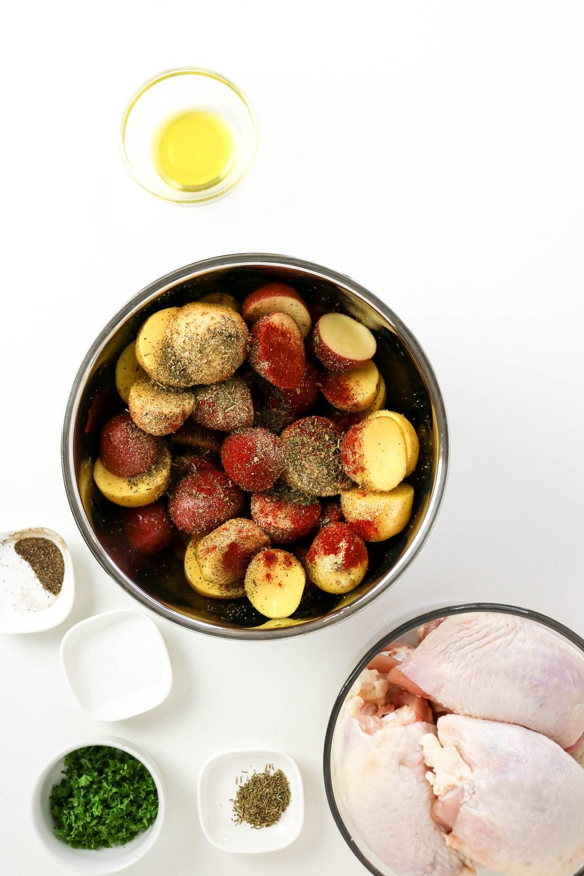 A metal bowl filled with halved potatoes seasoned with spices, next to raw chicken thighs and small bowls of oil, salt, pepper, parsley, and dried herbs.