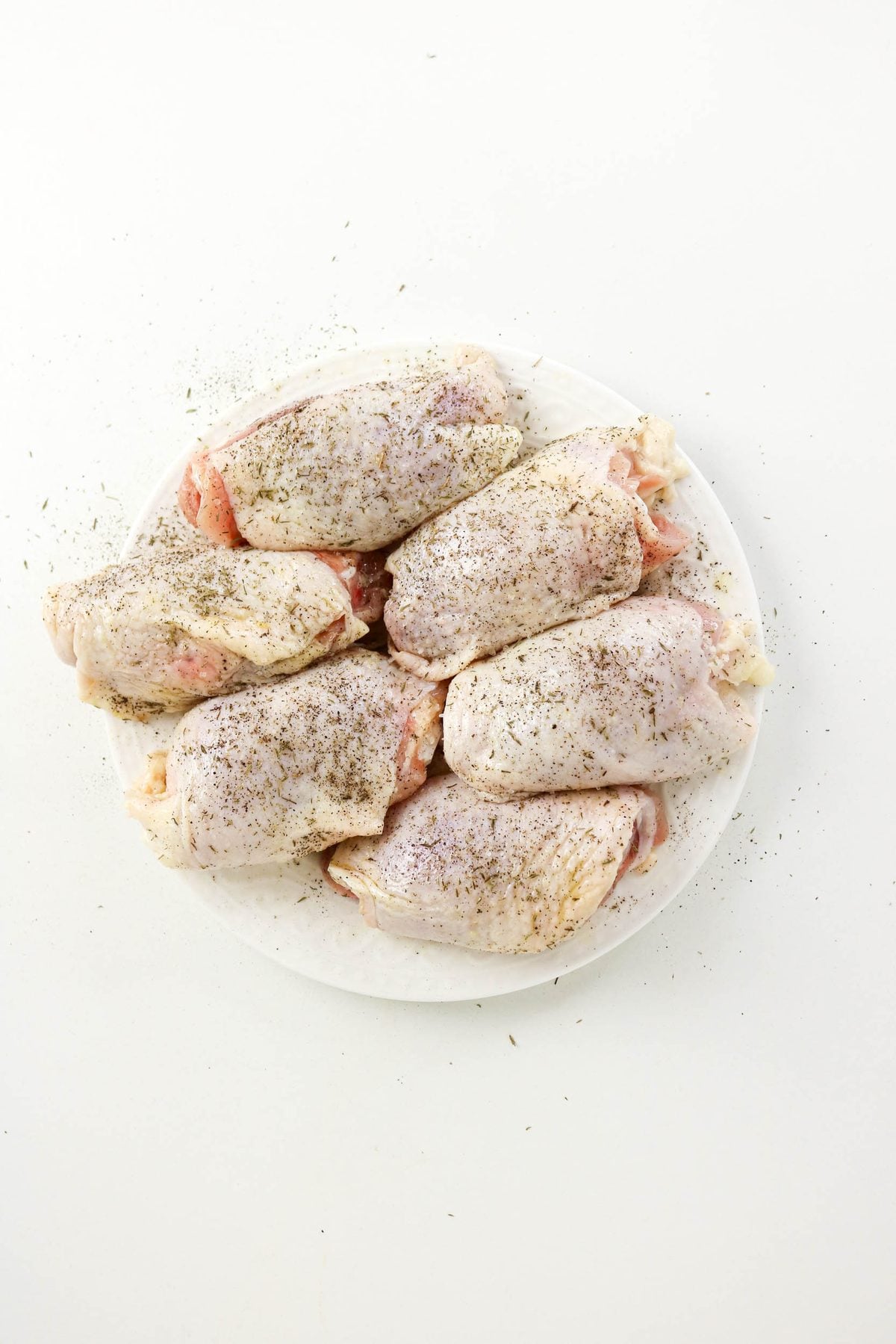 Six raw, skin-on chicken thighs seasoned with herbs on a white plate.