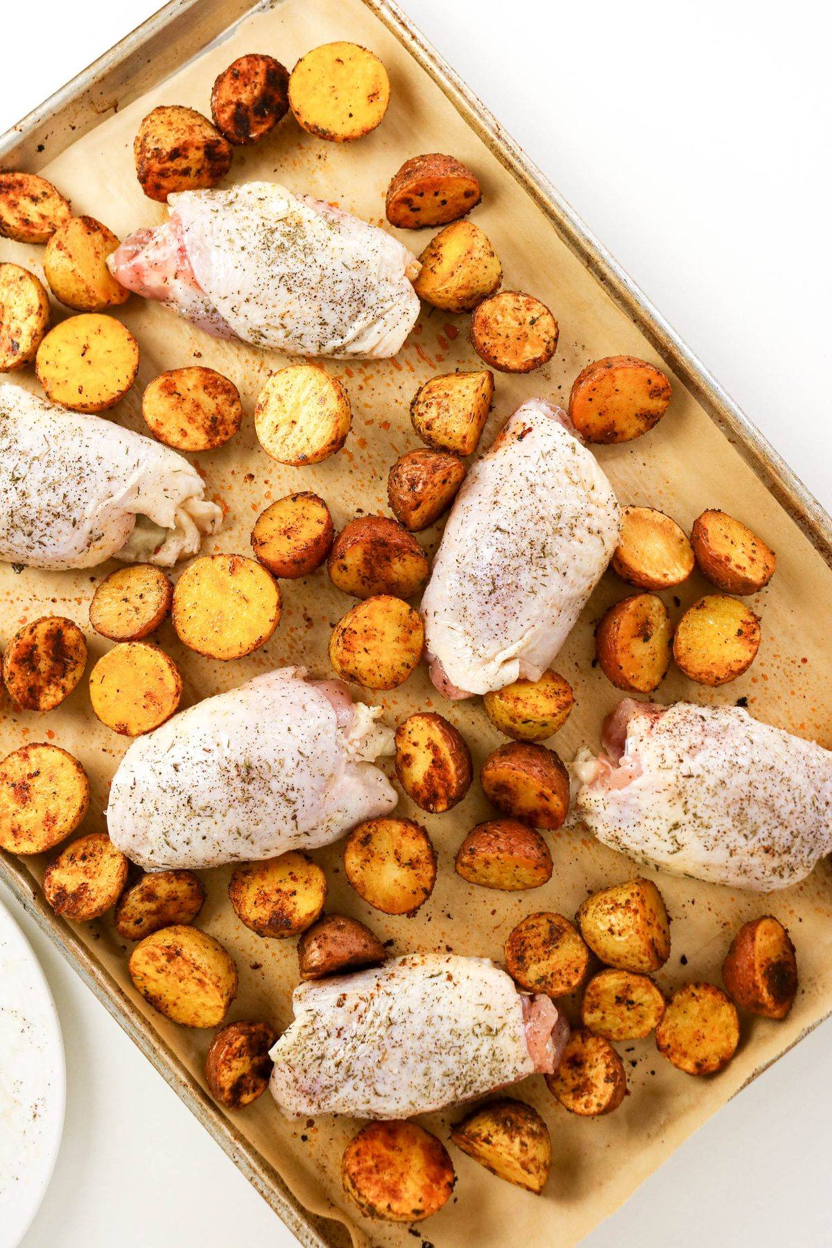 Raw chicken thighs and roasted potato wedges on a parchment-lined baking sheet.